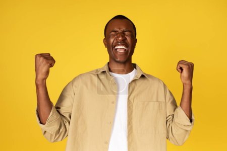 Foto de Feliz chico afroamericano joven en casual levanta los puños, se regocija por el éxito, celebra la victoria, aislado en el fondo del estudio amarillo. Ganador en la competencia, gran reacción de noticias, venta - Imagen libre de derechos