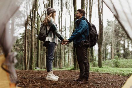 Photo for Happy young caucasian lady and man in jackets holding hands near tent, enjoy adventure, free time together at weekend, walk in cold forest in autumn. Love, relationship, vacation, active lifestyle - Royalty Free Image