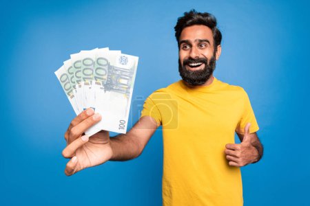 Foto de Emocionado indio rico de mediana edad hombre sosteniendo el dinero del euro y el gesto pulgar hacia arriba, celebrando la gran suerte financiera, posando sobre fondo azul. Concepto de beneficio y riqueza - Imagen libre de derechos
