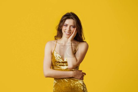 Photo for Gorgeous european lady posing wearing golden festive dress, smiling to camera over yellow studio background. Stylish woman expressing positive emotions - Royalty Free Image