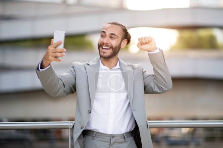 Téléchargez les photos : Jeune homme d'affaires excité célébrant le succès tout en regardant Smartphone en plein air, heureux entrepreneur masculin excité a de bonnes nouvelles, Réagissant émotionnellement à l'affaire réussie ou démarrage Bet, Espace libre - en image libre de droit