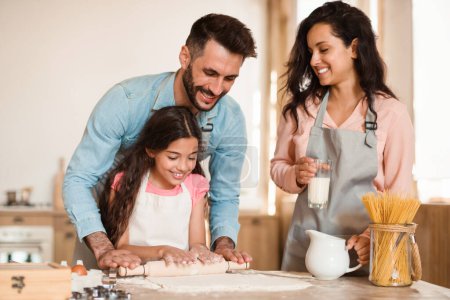Photo for Loving parents teaching their daughter baking pastry, child girl rolling out dough for cookies, family of three cooking together in kitchen. Weekend leisure concept - Royalty Free Image