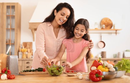 Foto de Mujer europea alegre abrazando a su hija disfrutando de cocinar ocio, hacer ensalada de verduras frescas en interiores, pasar tiempo en la cocina casera en interiores. Alimentación familiar y nutrición - Imagen libre de derechos