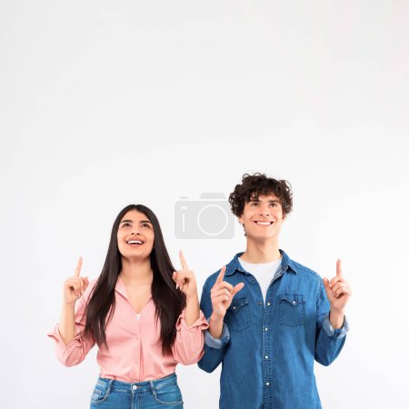 Photo for Look Up. Excited Young Couple Showing Free Space Pointing Fingers Up, Advertising Text Above Their Heads, Looking Upward While Standing Over White Studio Background. Square Shot - Royalty Free Image