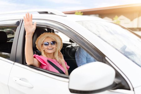 Téléchargez les photos : Joyeux belle femme âgée blonde agitant la fenêtre ouverte de la voiture et souriant à la caméra, portant des lunettes et un chapeau en osier. Touriste senior voyageant avec son mari en automobile - en image libre de droit