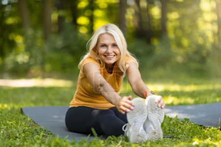 Fitness extérieur. Portrait d'une belle femme âgée étirant les muscles des jambes, dame mûre sportive assise sur un tapis de fitness sur la pelouse dans le parc, bénéficiant d'entraînements sportifs et d'un mode de vie actif, Copier l'espace