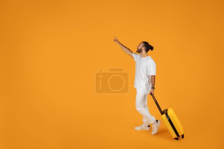 Foto de Sonriente sorprendido adulto europeo chico en camiseta blanca con la maleta señala el dedo en el espacio vacío, aislado en fondo de estudio naranja. Emociones del turismo, anuncio y oferta, viajes de vacaciones de verano - Imagen libre de derechos