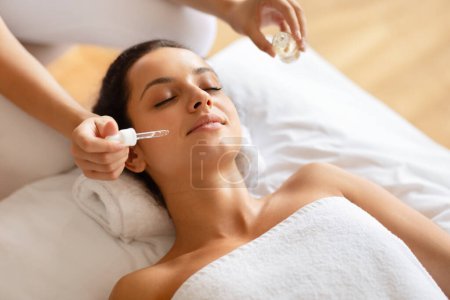 Photo for Facial Spa. Young Lady Enjoying Relaxing Face Massage While Masseur Applying Serum Moisturizing Skin At Luxury Spa Salon Interior, Woman Wrapped In Towel Closing Eyes During Skincare Treatment - Royalty Free Image