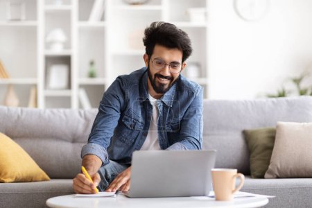 Photo for Online Education. Young smiling indian man using laptop and taking notes to notepad while sitting on couch at home, happy eastern guy watching webinar, study in internet, attending virtual lesson - Royalty Free Image