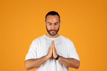 Photo for Serious adult european guy with beard in white t-shirt praying with hands gesture, isolated on orange studio background. Request, desire and faith, hope, good intentions sign - Royalty Free Image
