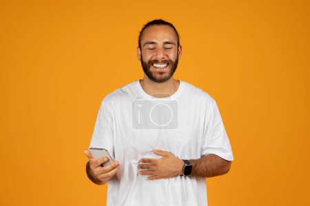 Foto de Riendo hombre europeo adulto con barba en camiseta blanca utiliza el teléfono para el chat, aislado en fondo de estudio naranja. Diversión con el dispositivo, emociones humanas, anuncio y oferta, aplicación de broma, buenas noticias - Imagen libre de derechos