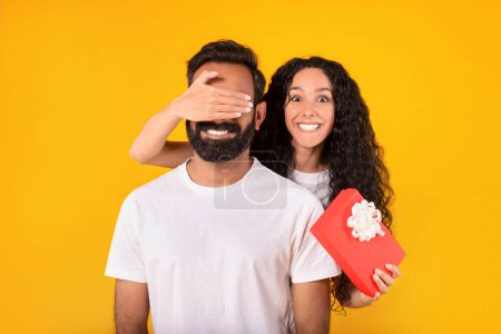 Photo for Loving middle eastern woman closing mans eyes from back holding present box, surprising husband on valentines day with romantic gift over yellow background. Anniversary celebration and presents - Royalty Free Image