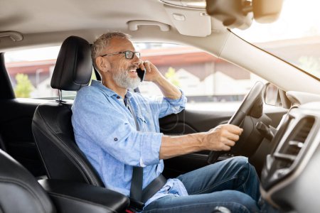 Photo for Cheerful handsome bearded elderly caucasian man driver talking on phone while driving car, looking at road and smiling. Happy gentleman have conversation on cell phone, going to office by auto - Royalty Free Image