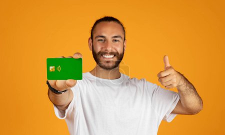 Foto de Sonriente adulto europeo chico en camiseta blanca muestran tarjeta de crédito y pulgar hacia arriba aislado en fondo de estudio naranja. Crédito de aprobación, beneficio financiero de recomendación, ahorro, asesoramiento profesional - Imagen libre de derechos