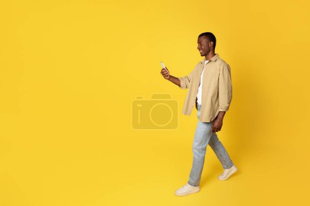 Photo for Positive millennial african american man in casual use phone for chat, walk, isolated on yellow studio background, full length. Spare time, app, social networks, blog, good news - Royalty Free Image
