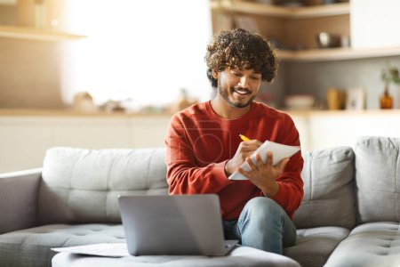 Photo for Online Education. Smiling Indian Male Study With Laptop At Home And Taking Notes, Happy Millennial Eastern Man Using Computer And Writing In Notepad, Enjoying Remote Study, Copy Space - Royalty Free Image
