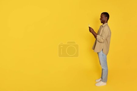 Photo for Cheerful millennial african american guy in casual chatting on phone, isolated on yellow studio background, full length, profile. Ad and offer, communication remotely app, social networks, blog - Royalty Free Image