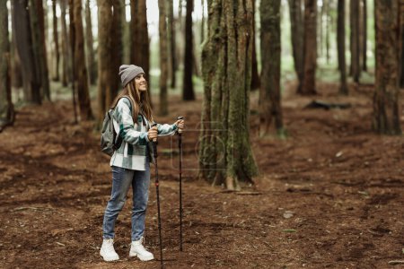 Photo for Positive young caucasian woman in jacket, hat with trekking sticks walk, enjoy adventure, free time at weekend in cold forest in autumn. Vacation, travel alone, active lifestyle - Royalty Free Image