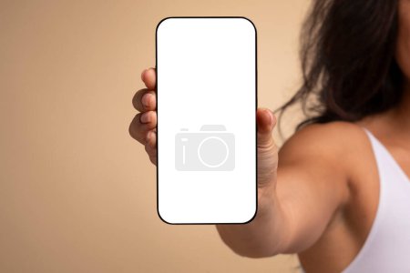 Foto de Irreconocible morena de pelo largo mujer con top blanco sosteniendo en su mano, mostrando teléfono inteligente con blanco pantalla en blanco maqueta en beige fondo de estudio, increíble oferta en línea, aplicación móvil - Imagen libre de derechos