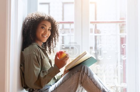 Photo for Smiling black young lady relaxing on windowsill with a paper book and holding apple, enjoying her bookworm weekend at home, smiling to camera. Happy woman spending free time to relax with a novel - Royalty Free Image