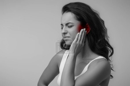 Photo for Black and white photo of young brunette eastern lady suffering from otitis, rubbing her inflamed ear highlighted with red over grey background, ear diseases concept, copy space - Royalty Free Image