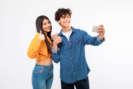 Téléchargez les photos : On l'aime bien. Joyeux jeune couple adolescent posant pour Selfie, prenant des photos sur Smartphone et geste pouces vers le haut dans l'approbation sur fond blanc, Studio Shot. Médias sociaux et gadgets - en image libre de droit