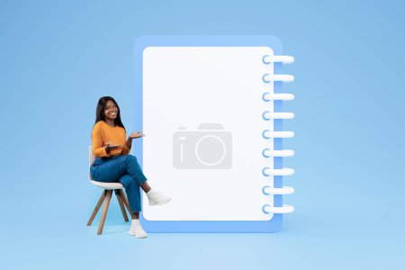 Photo for Positive young black woman in casual sit on chair point hands on big plan list with free space, isolated on blue studio background. Day planning, motivation, checklist recommendation - Royalty Free Image