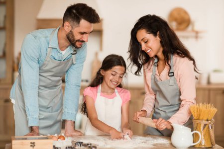 Photo for Cheerful parents and their daughter baking pastry in kitchen together, child girl kneading dough on table while preparing cookies or pizza, family enjoying cooking homemade food - Royalty Free Image