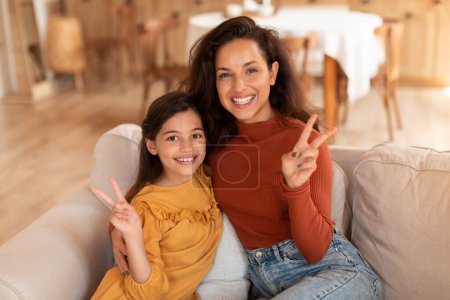 Foto de Señal de victoria. Sonriendo mamá e hija mostrando el gesto de paz divirtiéndose posando juntas en el sofá en casa, sentadas y abrazando el gesto V-Sign to Camera. Madre y niño chica vínculo - Imagen libre de derechos