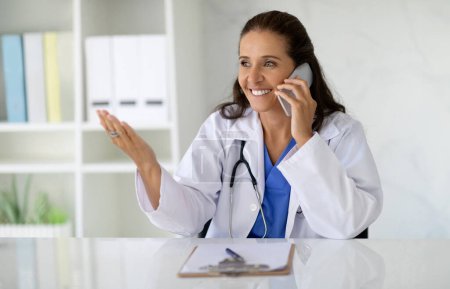 Téléchargez les photos : Joyeux attrayant brunette caucasienne mature femme médecin assis au bureau, parlant au téléphone, souriant et gestuel, consultant patient, avoir rendez-vous téléphonique, copier l'espace - en image libre de droit