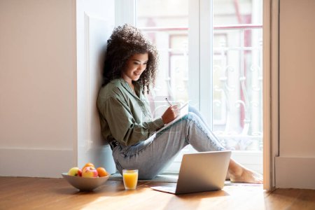 Photo for E-Learning. Young brazilian lady writing in notebook looking at laptop while working and studying remotely indoor, sitting on floor near window at home. Bushy woman student taking notes near pc - Royalty Free Image