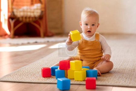 Téléchargez les photos : Concept de jouets de développement. Bébé bébé mignon jouant avec des blocs de construction colorés à la maison, adorable petit garçon ou fille portant une combinaison de corps assis sur le tapis dans le salon, Copier l'espace - en image libre de droit