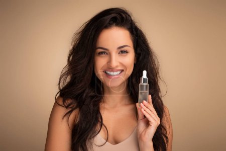 Photo for Skin hydration, anti-aging. Portrait of cheerful millennial brunette long-haired lady with glowing flawless skin holding bottle with moisturizing serum or oil for face, beige studio background - Royalty Free Image