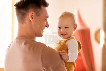 Foto de Retrato de feliz padre joven sosteniendo a su adorable hijo bebé o hija en las manos y sonriendo, alegre papá milenario vinculándose con lindo niño en casa, disfrutando del tiempo con el niño - Imagen libre de derechos