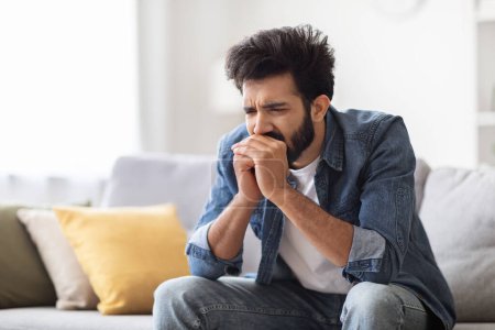 Photo for Portrait of stressed depressed young indian man sitting on couch at home, worried eastern male covering his mouth with hands, thinking about something, having life problems, copy space - Royalty Free Image