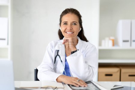 Photo for E-medicine, telehealth concept. Cheerful middle aged woman wearing medical coat doctor sitting at desk at clinic, using wireless earphones earbuds, doc have online consultation with patient - Royalty Free Image
