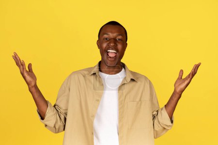 Photo for Glad millennial african american man in casual raises hands, rejoices to success, celebrates win, isolated on yellow studio background. Victory, great news, huge sale gesture - Royalty Free Image