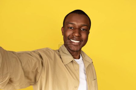 Photo for Smiling millennial african american man in casual taking selfie, shooting video blog, isolated on yellow studio background. Ad and offer, recommendation app, video call, photo for social networks - Royalty Free Image