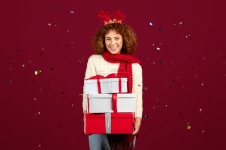 Téléchargez les photos : Joyeux adolescent caucasien fille bouclée avec bois de cerf et écharpe tenir beaucoup de boîtes avec cadeau et confettis, isolé sur fond de studio rouge. Mode de vie, Nouvel An, Fête de Noël - en image libre de droit