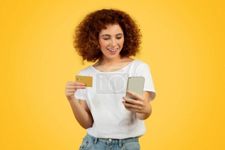 Photo for Cheerful teenage caucasian curly girl with credit card pays with phone for order, isolated on orange studio background. Fashion lifestyle, sale, online shopping and banking, ad and offer - Royalty Free Image