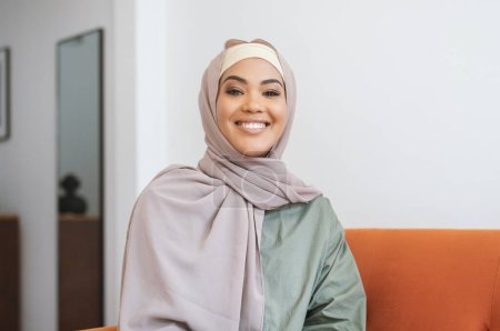 Téléchargez les photos : Portrait de femme musulmane heureuse dans le hijab posant à l'intérieur de la maison, portant des vêtements décontractés et souriant à la caméra assis sur un canapé confortable dans le salon moderne. Concept de beauté féminine - en image libre de droit