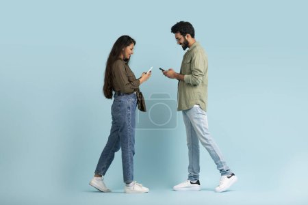 Photo for Indian millennial man and woman with phones in their hands walking towards each other, chatting with friends or texting one another, blue background. Communication, virtual world concept - Royalty Free Image