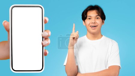 Photo for Positive young asian guy pointing finger up, got idea, near hand with phone with empty screen, isolated on blue background, studio, collage. Solution, brainstorm, app recommendation - Royalty Free Image