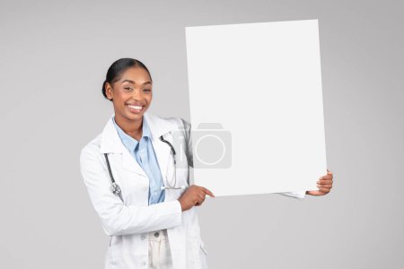 Photo for Cheerful black woman doctor in white coat confidently holding blank banner, isolated on gray background studio, expressing positivity. Healthcare services in clinic, ad and offer - Royalty Free Image
