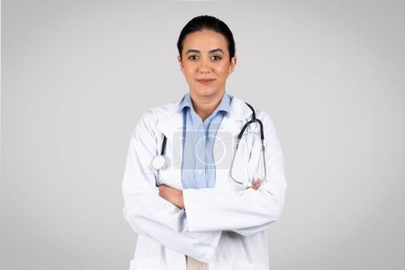 Téléchargez les photos : Portrait d'une femme médecin latino confiante aux bras croisés, souriante à la caméra, debout sur fond de studio gris. Mettre en valeur la personnalité professionnelle des soins de santé - en image libre de droit