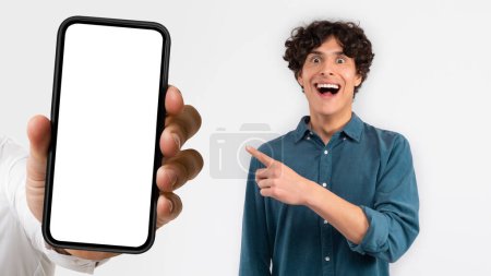 Photo for Glad shocked surprised millennial european man pointing finger at hand with smartphone with empty screen, isolated on white background, studio, collage. Fun recommend, joke, app, website - Royalty Free Image