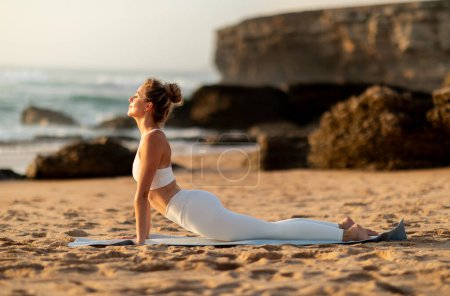 Téléchargez les photos : Jeune femme mince européenne calme pratiquant le yoga à asana, faisant de l'exercice à l'extérieur sur la plage de la mer, vue latérale. Stretching, sport, entraînement fitness, soins de santé et pilates - en image libre de droit