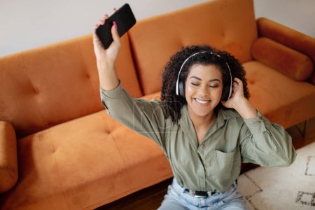 Téléchargez les photos : High Angle Of Smiling Relaxed Black Lady Écouter de la musique sur smartphone portant des écouteurs sans fil, lever la main avec le téléphone tout en appréciant la chanson préférée à la maison, Focus sélectif - en image libre de droit