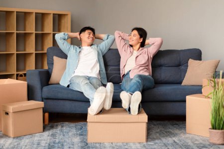 Foto de Oferta inmobiliaria. La feliz pareja asiática hace una pausa reparadora apoyándose en el sofá moviendo un nuevo apartamento de alquiler, relajándose en interiores entre cajas de cartón, compartiendo un momento de felicidad - Imagen libre de derechos