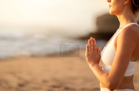 Téléchargez les photos : Jeune femme méconnaissable méditant seule sur la plage de l'océan, tenant la main dans un geste namaste, pratiquant le yoga à l'extérieur, vue latérale, espace de copie, recadré - en image libre de droit